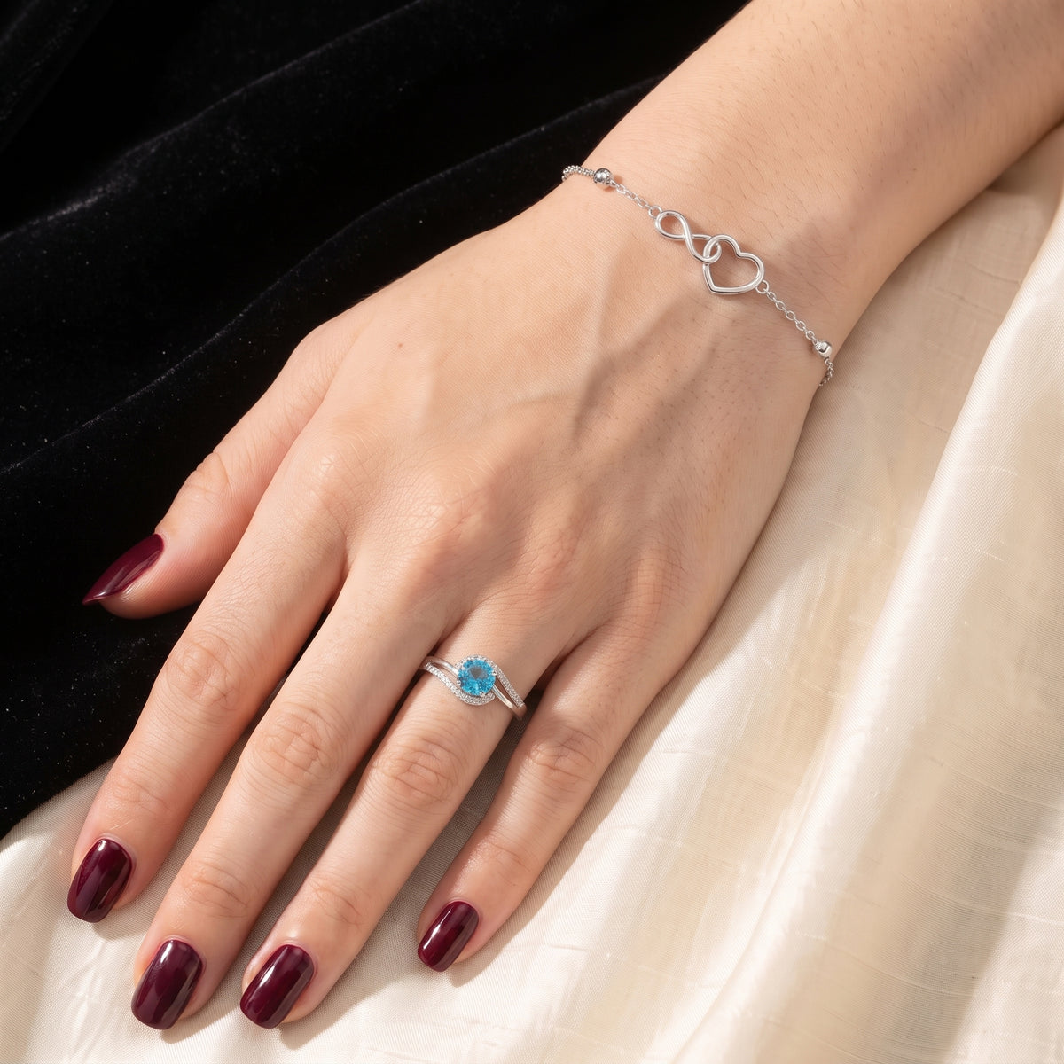 Bransoletka na opartej dłoni z bordowym manicure, w kadrze turkusowy pierścionek i jasna jedwabna tkanina